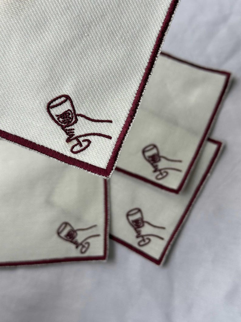 Embroidered white cotton cocktail coasters with burgundy stitching of an embroidered hand holding a wine glass