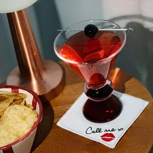 K&ouml;nnte beinhalten: Ein roter Cocktail in einem Martini-Glas mit Stiel, garniert mit einer schwarzen Kirsche, auf einem wei&szlig;en quadratischen Untersetzer mit der Aufschrift "Call me xo" und einem roten Lippen-Design. Eine Sch&uuml;ssel mit Chips und eine Kupferlampe sind ebenfalls zu sehen.