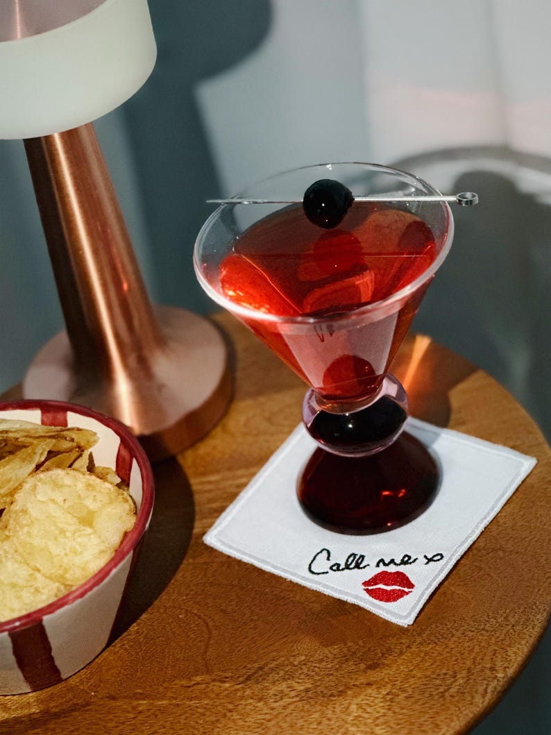 K&ouml;nnte beinhalten: Ein roter Cocktail in einem Martini-Glas mit Stiel, garniert mit einer schwarzen Kirsche, auf einem wei&szlig;en quadratischen Untersetzer mit der Aufschrift "Call me xo" und einem roten Lippen-Design. Eine Sch&uuml;ssel mit Chips und eine Kupferlampe sind ebenfalls zu sehen.