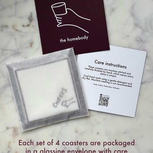 K&ouml;nnte beinhalten: Ein Set Untersetzer, verpackt in einem Pergamentumschlag mit Pflegehinweisen. Die Untersetzer sind wei&szlig; und tragen das Wort "CHEERS". Die Verpackung enth&auml;lt eine Karte mit den Worten "the homebody" und Pflegehinweisen.