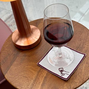 A glass of wine on a cotton coaster with an embroidered hand holding a wine glass in a raised cheers position