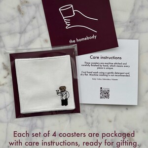May include: A set of four white cotton coasters, each with an embroidered bear design, packaged with care instructions. The coasters are presented with a burgundy card featuring a hand holding a cup and the text "the homebody".