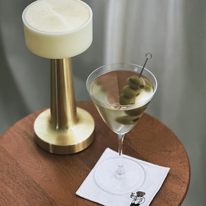May include: A martini in a stemmed glass with olives on a pick, resting on a white square coaster with a bear illustration. A gold-colored lamp with a cream-colored top sits on a wooden table.