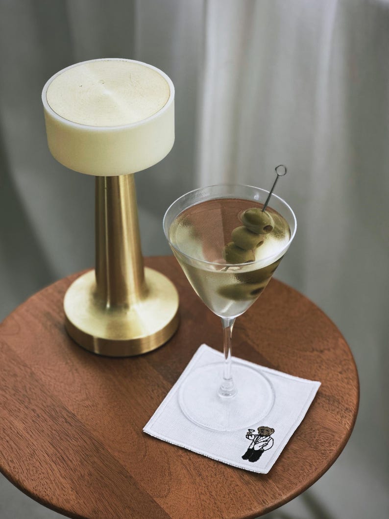 May include: A martini in a stemmed glass with olives on a pick, resting on a white square coaster with a bear illustration. A gold-colored lamp with a cream-colored top sits on a wooden table.