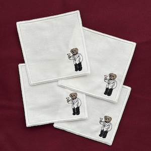 May include: Four white square coasters with embroidered images of a bear wearing a suit and holding a cocktail glass. The coasters have a white stitched border and are arranged on a burgundy surface.