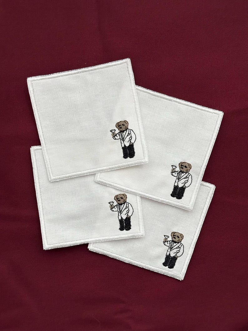 May include: Four white square coasters with embroidered images of a bear wearing a suit and holding a cocktail glass. The coasters have a white stitched border and are arranged on a burgundy surface.