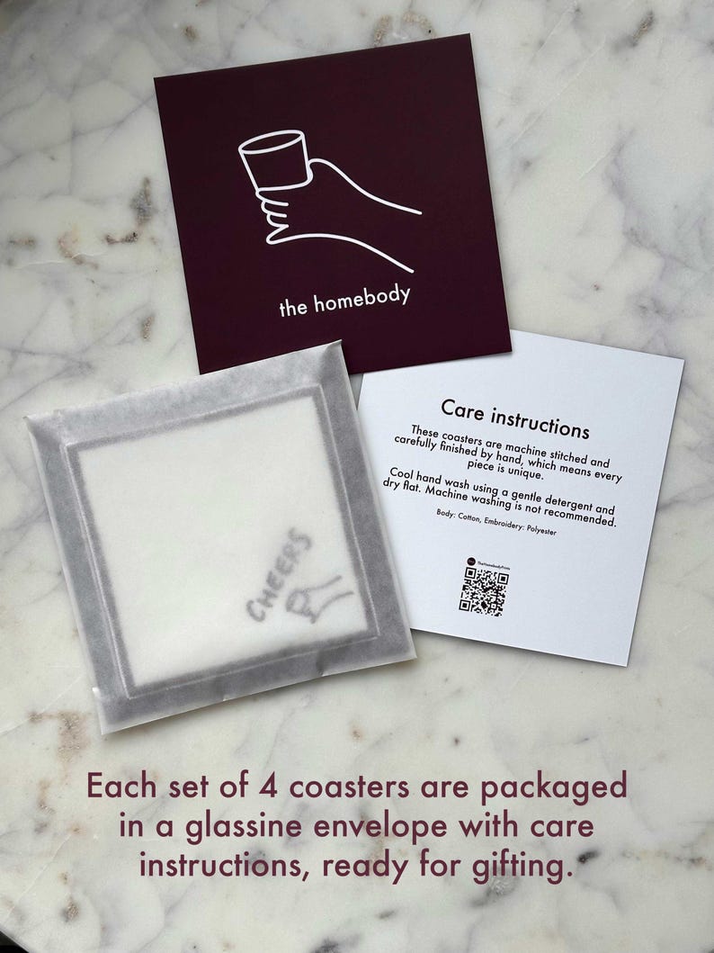 May include: Set of four coasters packaged in a clear glassine envelope with care instructions. The coasters feature a white "Cheers" graphic on a grey background. The envelope has a burgundy card with a white illustration of a hand holding a glass and the text "the homebody".