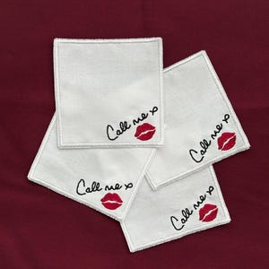 K&ouml;nnte beinhalten: Vier wei&szlig;e quadratische Untersetzer mit gesticktem Text und einem roten Lippen-Design. Der Text lautet "Call me xo" in schwarzer Schrift. Die Untersetzer sind auf einer dunkelroten Oberfl&auml;che angeordnet.
