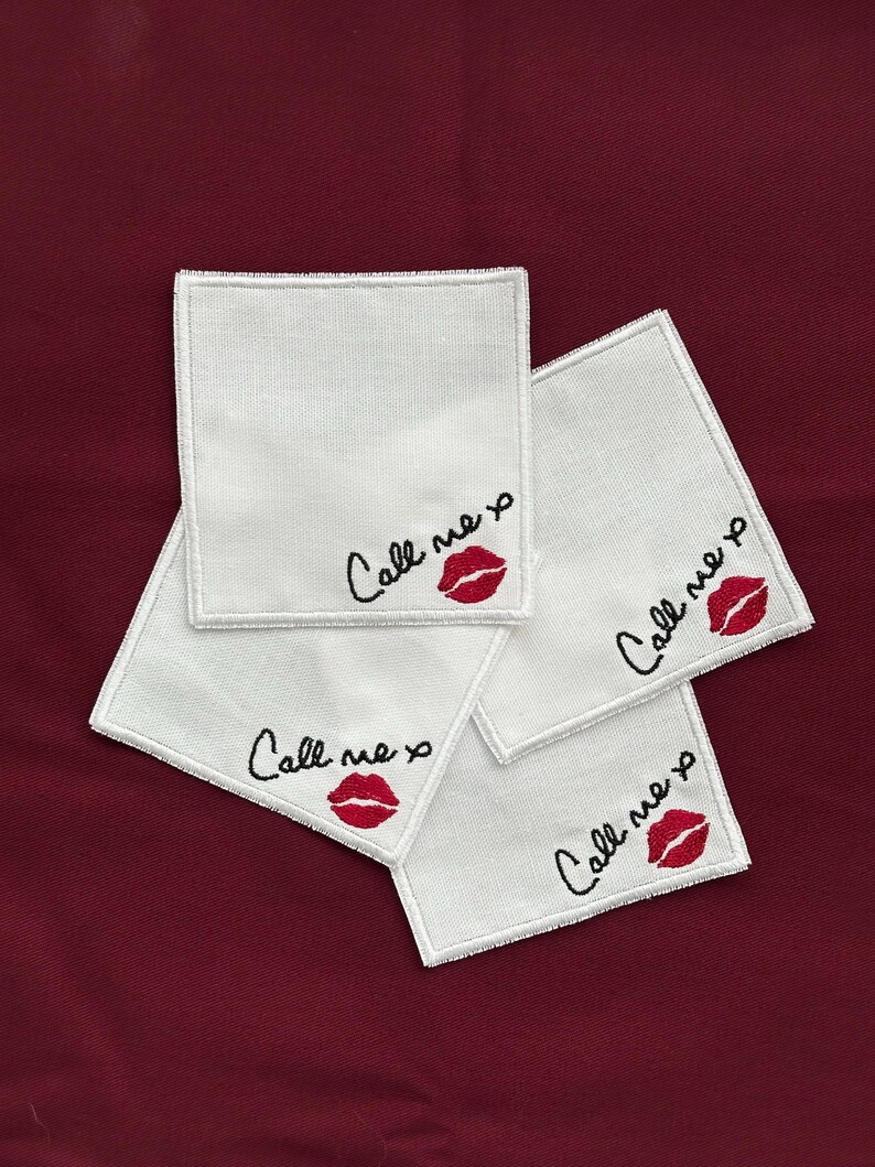 K&ouml;nnte beinhalten: Vier wei&szlig;e quadratische Untersetzer mit gesticktem Text und einem roten Lippen-Design. Der Text lautet "Call me xo" in schwarzer Schrift. Die Untersetzer sind auf einer dunkelroten Oberfl&auml;che angeordnet.