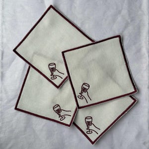 Four embroidered white cotton cocktail coasters with burgundy stitching of an embroidered hand holding a wine glass