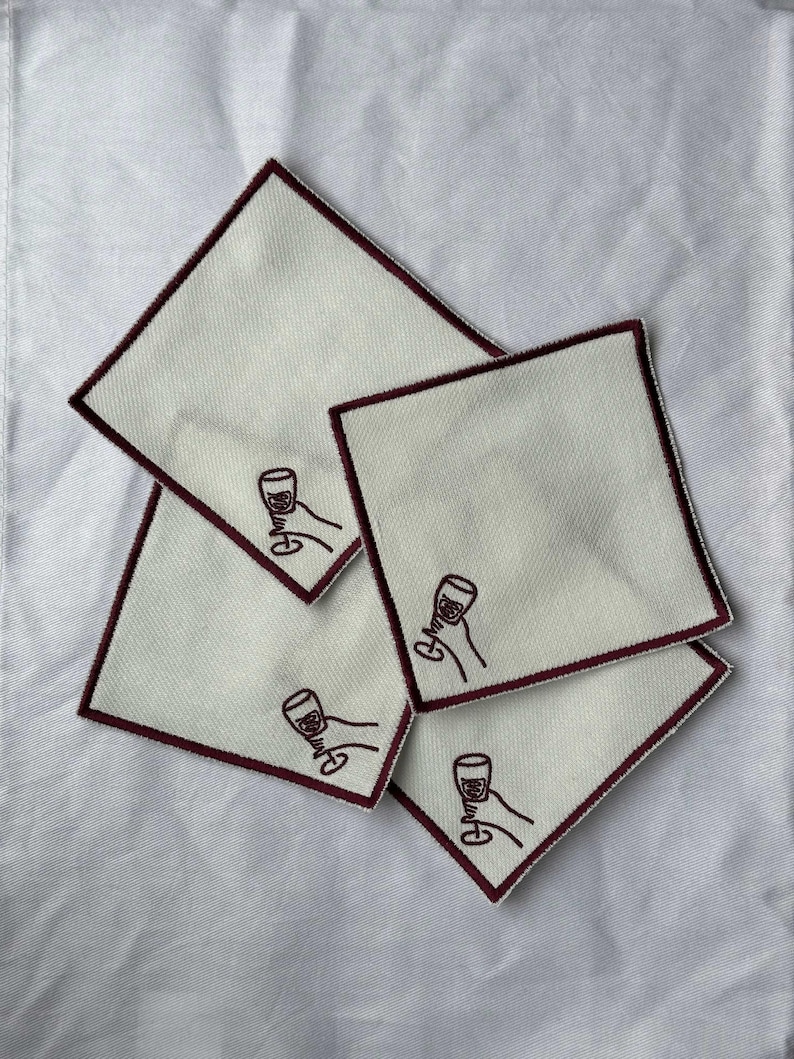 Four embroidered white cotton cocktail coasters with burgundy stitching of an embroidered hand holding a wine glass