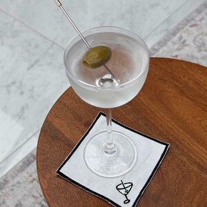 A vodka martini with an olive garnish on an embroidered cocktail napkin which has a stitched martini glass motif