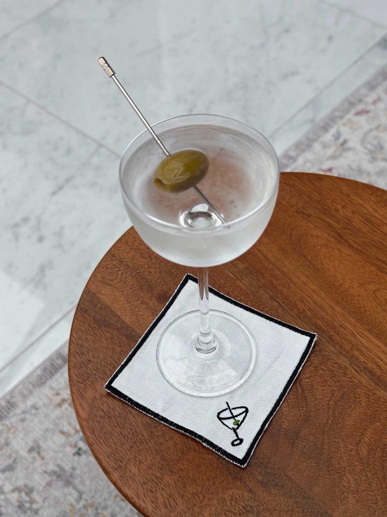 A vodka martini with an olive garnish on an embroidered cocktail napkin which has a stitched martini glass motif