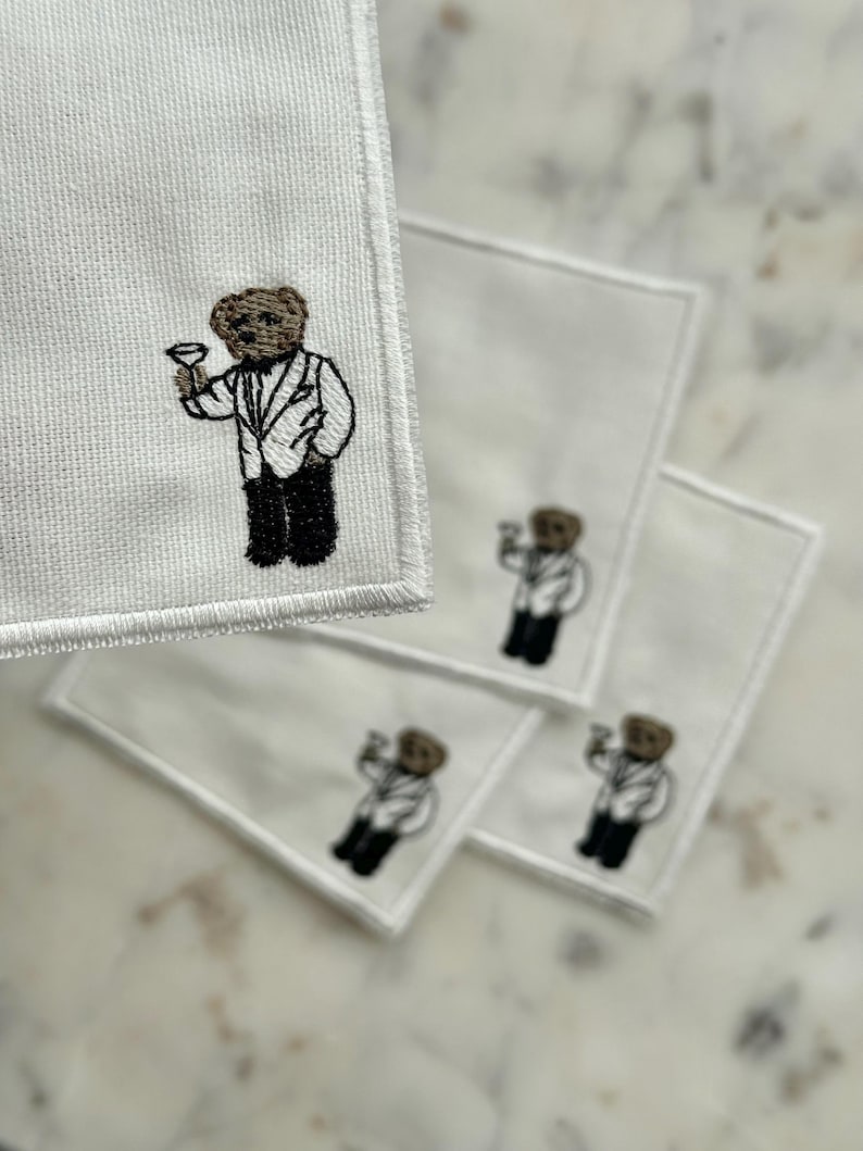 May include: White square cocktail napkins with an embroidered brown bear wearing a tuxedo and holding a martini glass. The bear is the focal point of the design, repeated on multiple napkins.