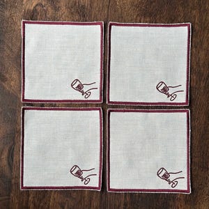 Four embroidered white cotton cocktail coasters with burgundy stitching of an embroidered hand holding a wine glass, on a wooden background