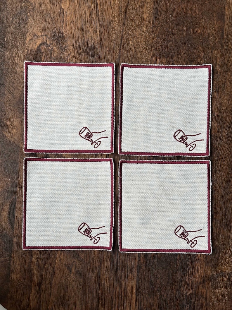 Four embroidered white cotton cocktail coasters with burgundy stitching of an embroidered hand holding a wine glass, on a wooden background
