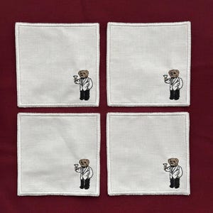 May include: Four white square coasters with a stitched design of a bear wearing a tuxedo and holding a cocktail glass. The coasters are arranged on a burgundy surface.