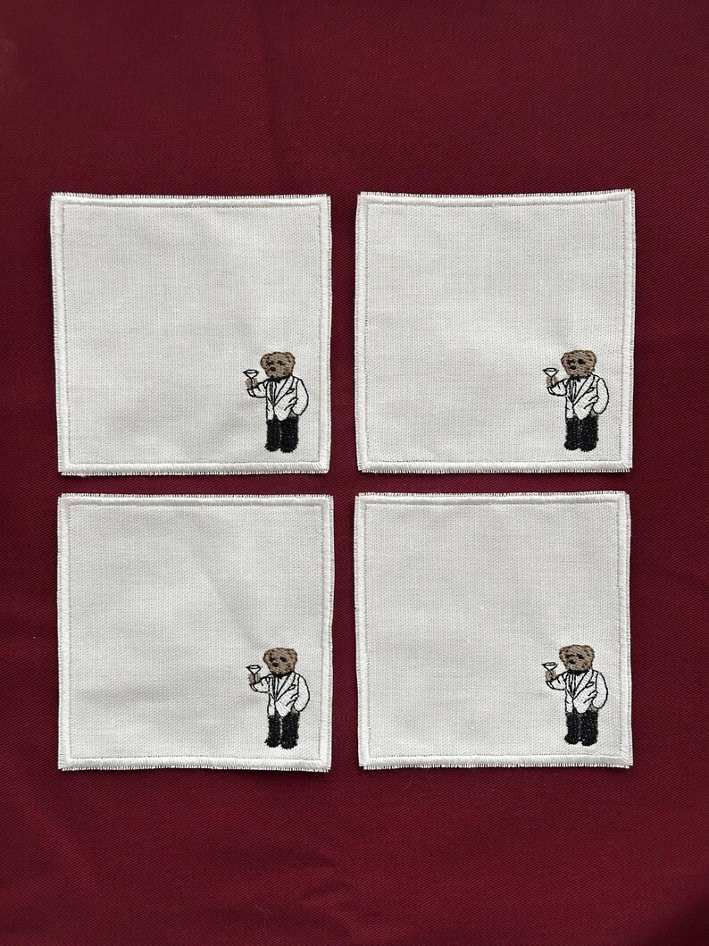 May include: Four white square coasters with a stitched design of a bear wearing a tuxedo and holding a cocktail glass. The coasters are arranged on a burgundy surface.
