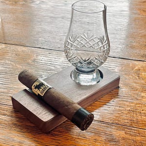 May include: A clear crystal whiskey glass with a diamond pattern sits on a wooden coaster next to a cigar. The cigar has a black band with gold lettering and a bull logo. The coaster is made of dark wood.