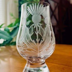 May include: A clear glass tumbler with a curved shape and a thick base, featuring an etched thistle design on the front. The base of the glass has the text The Glencairn Glass etched into the bottom, visible through the clear glass material.