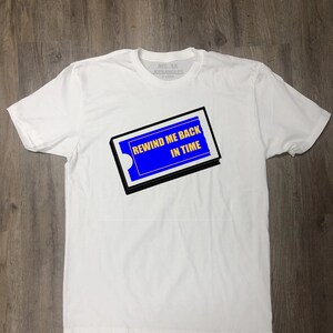 May include: White t-shirt with a blue and black graphic of a VHS tape with the text "REWIND ME BACK IN TIME".