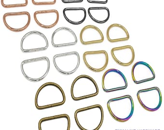 May include: Assortment of metal D-rings in various finishes, including silver, gold, bronze, copper, black, and rainbow. The D-rings are arranged in rows, with each row featuring a different finish. The D-rings are all the same size and shape.