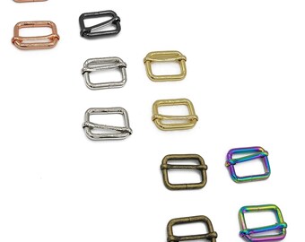 Op de afbeelding: Assortiment van metalen schuifgespen in verschillende afwerkingen, waaronder zilver, goud, zwart, brons en regenboog. De gespen zijn allemaal dezelfde maat en vorm, met een rechthoekig ontwerp en een kleine opening aan elke kant.