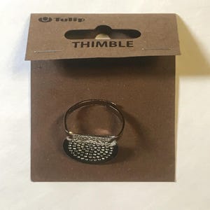 May include: A silver metal thimble with a textured surface. The thimble is on a brown cardboard backing with the word "THIMBLE" printed on it.