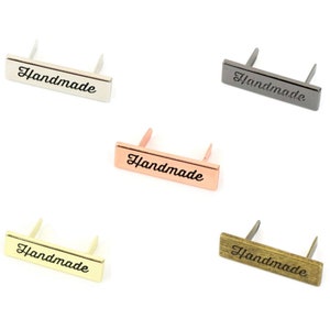 Może przedstawiać: Pięć metalowych etykiet z wygrawerowanym napisem "Handmade". Etykiety są w różnych kolorach: srebrny, czarny, różowe złoto, złoty i brązowy.