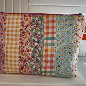 May include: A patchwork cosmetic bag with a variety of floral and gingham patterns in pink, yellow, blue, and red. The bag has a pink zipper and a small pink and white checkered fabric loop on the side. An orange tag is attached.