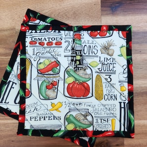 May include: Two quilted pot holders with a black border and a white background. The pot holders feature a colorful print of tomatoes, peppers, onions, and other ingredients in jars. The text includes measurements and cooking terms.