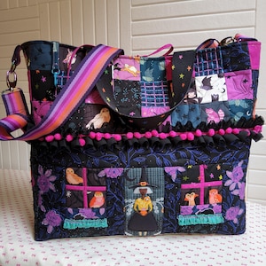 May include: A quilted tote bag featuring a patchwork design with Halloween-themed motifs including owls, a witch, and stars. The bag has a black base with purple and teal accents, a pink pom-pom trim, and a striped purple and orange shoulder strap.