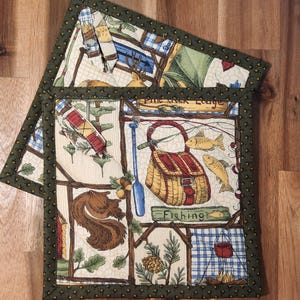 May include: Two green-trimmed pot holders with a woodland theme. The pot holders feature a fishing scene with a basket, a paddle, and fish. The text "Pine Creek Lodge" and "Fishing" are visible on the pot holders.