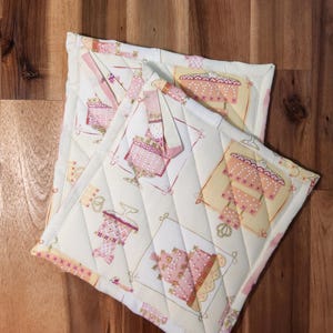 May include: Two quilted pot holders with a white background and pink and yellow cake designs. The pot holders have a quilted diamond pattern.