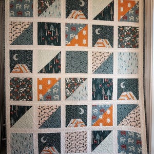 May include: A patchwork quilt with a repeating pattern of squares and triangles. The squares feature various nature-themed designs, including deer, trees, mountains, and moons. The color palette includes shades of green, orange, and cream, creating a cozy, rustic aesthetic.