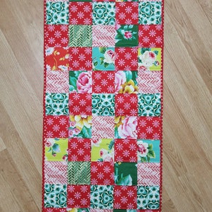 May include: A red, green, and white patchwork table runner with a floral pattern. The runner is made of fabric squares sewn together.