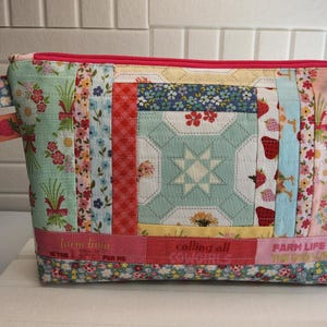 May include: A quilted fabric cosmetic bag with a pink zipper and a green zipper pull. The bag features a patchwork design with floral, strawberry, and star patterns. The bottom has text that reads "Farm Life is the life for me."