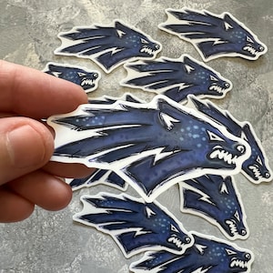 Puede incluir: Varias pegatinas de cabeza de lobo azules y blancas con un diseño estilizado. Las cabezas de lobo miran hacia la derecha y tienen una expresión feroz.