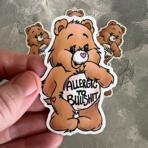 May include: A brown bear cartoon sticker with a pink heart on its chest and the text "Allergic to Bullshit" on its belly. The sticker is on a gray background.
