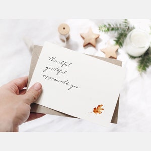 May include: A white card with the words "thankful grateful appreciate you" written in black cursive. A small, dried, orange leaf is in the bottom right corner of the card.