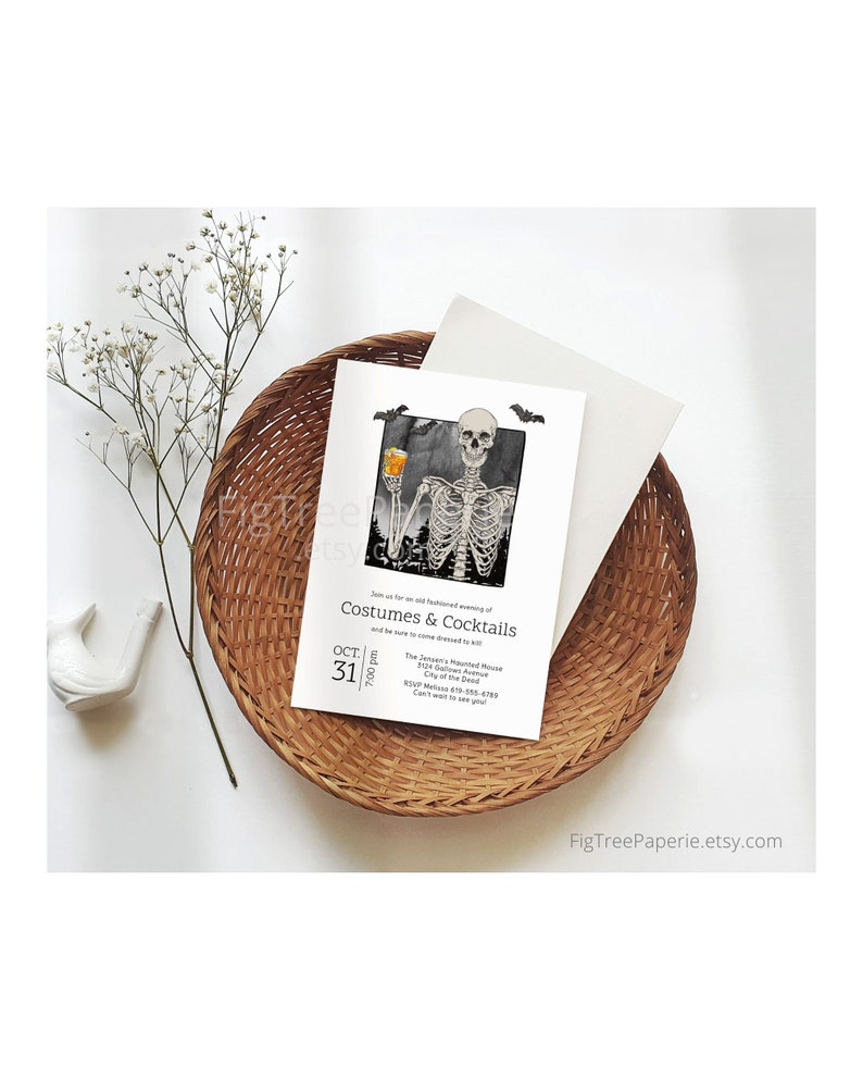 May include: Halloween-themed invitation with a skeleton holding a drink, set against a black and white background. The invitation is on a white card, with the text "Costumes & Cocktails" and event details. The card is placed in a woven basket.