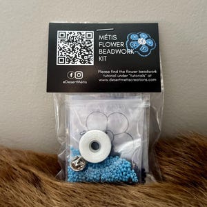 May include: A clear plastic bag containing a Métis Flower Beadwork Kit. The kit includes blue beads, white thread, and a silver pin. A black and white label with the text "MÉTIS FLOWER BEADWORK KIT" is attached to the bag.