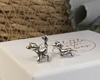May include: A pair of silver-toned deer stud earrings. The earrings are on a white box with the words "little birch paper" in a circular logo. The background includes a woven basket and a soft cloth.