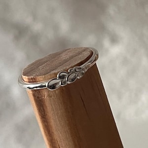 May include: A thin silver ring featuring a small, delicate cluster of rounded, petal-like shapes on the band. The ring is positioned horizontally around the top edge of a smooth, cylindrical wooden dowel, showcasing its polished metallic finish.