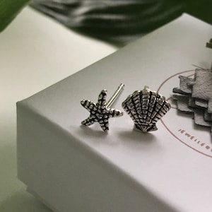 May include: A pair of silver stud earrings, one starfish and one seashell, presented on a white box. The earrings have textured surfaces. A green leaf is visible in the background.