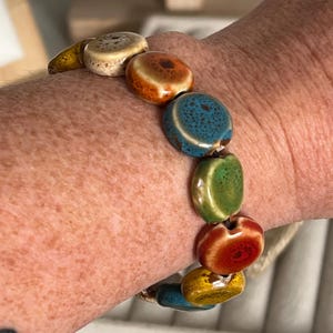 May include: A colorful ceramic bracelet with round, glazed beads in shades of beige, orange, teal, green, red, and yellow. The bracelet is strung on a neutral-colored cord and is worn on a wrist.