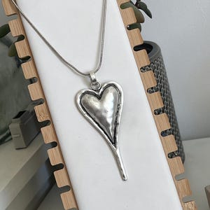 May include: A silver-colored heart pendant necklace. The pendant has a textured surface and a long, slender bottom. The necklace is displayed on a white surface, held by a wooden frame. The background includes a plant and a decorative pot.