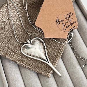 May include: A silver heart pendant necklace with a long, slender stem. The necklace is displayed on a beige burlap pouch, with a brown tag that reads "The Little Earth Comber". The necklace is displayed on a grey velvet surface.