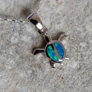 Puede incluir: Collar con colgante de tortuga plateada. El colgante de tortuga presenta una incrustación de ópalo azul y verde. La cadena del collar también es plateada. El colgante mide aproximadamente 2,5 cm de largo.