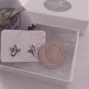 May include: Two silver angel earrings in a white box with a white card that says "The Little Angel Co.". A British one pound coin is next to the earrings for size comparison.